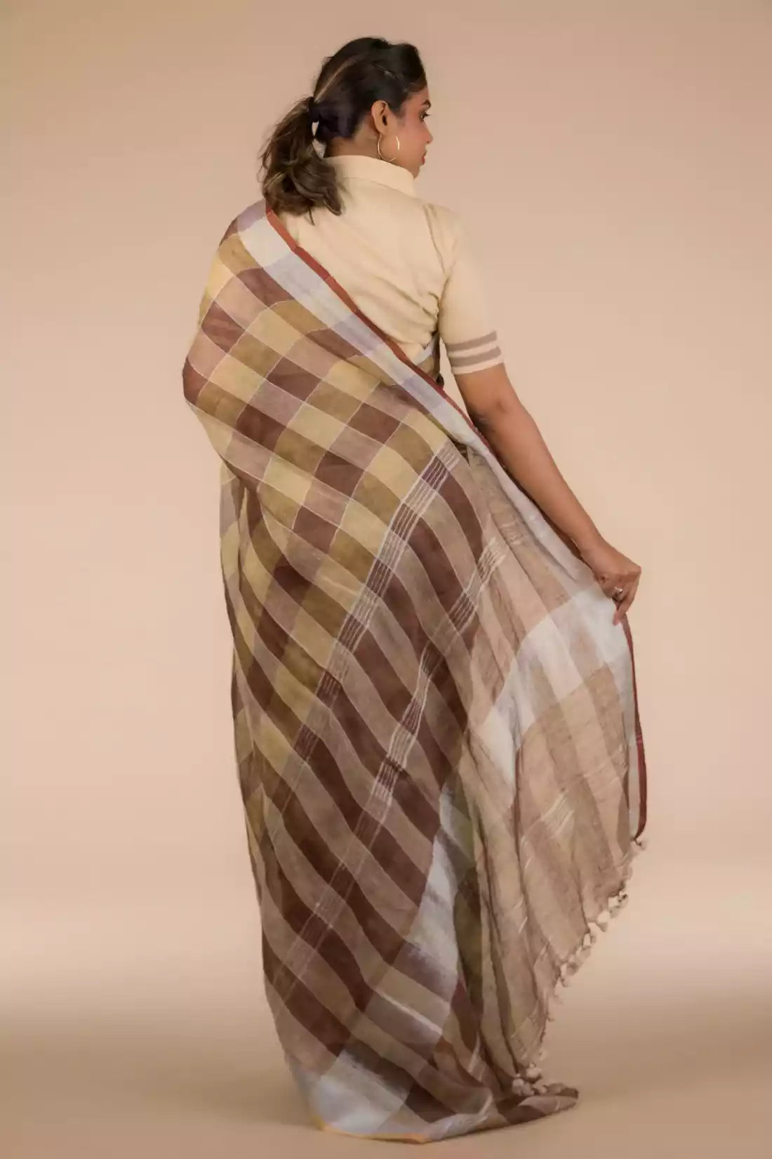 back view of a woman in ethnic posing wearing brown & beige checks saree with tie top blouse, flaunting loose end of her saree