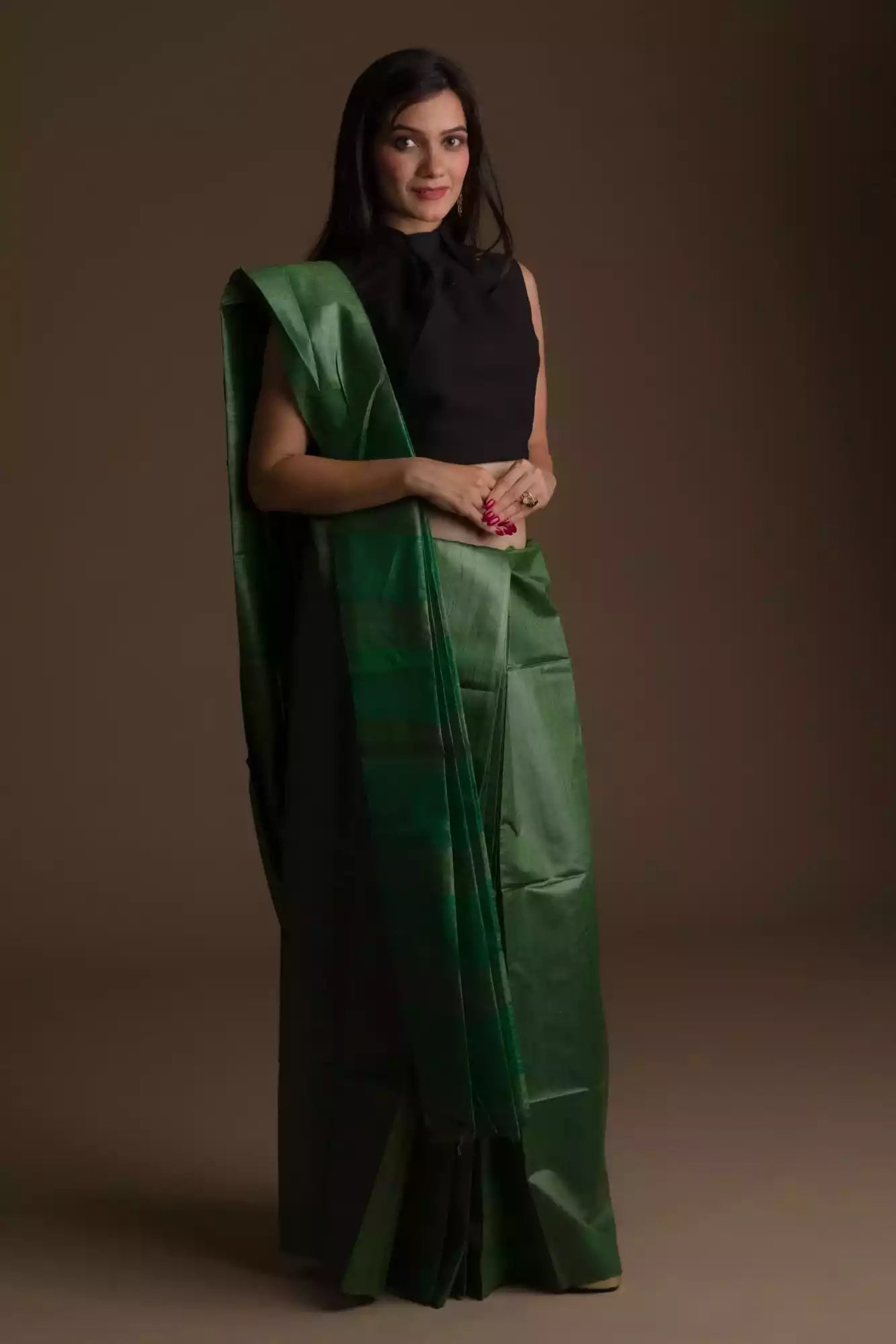 woman in ethnic posing wearing dark green saree with black blouse and heels against a beige background
