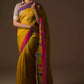 A lady in mustard with blue and pink border Jamdani hand weaving Saree standing against a brown background.
