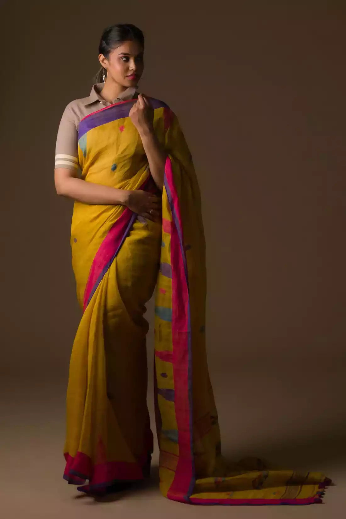 A lady in mustard with blue and pink border Jamdani hand weaving Saree standing against a brown background.