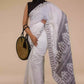 A lady in  Baluchari Resham Thread Linen Saree in White, womens workwear standing against a beige background looking sideways