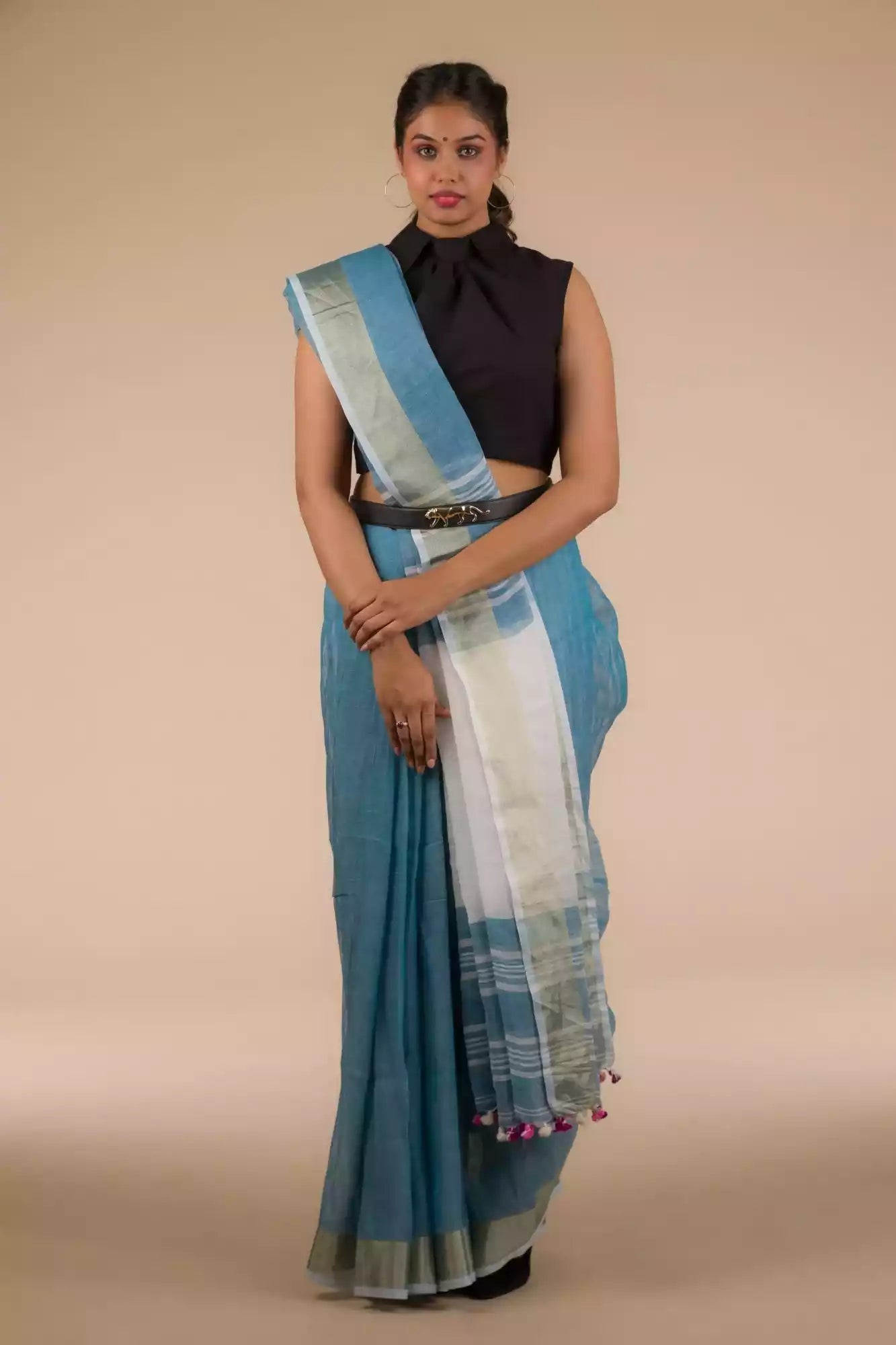 front view of a woman wearing a blue saree and a black blouse with stripes at the border and a belt in front of a beige backdrop