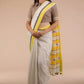 Front view of a woman in a white and yellow border saree standing in front of a beige backdrop