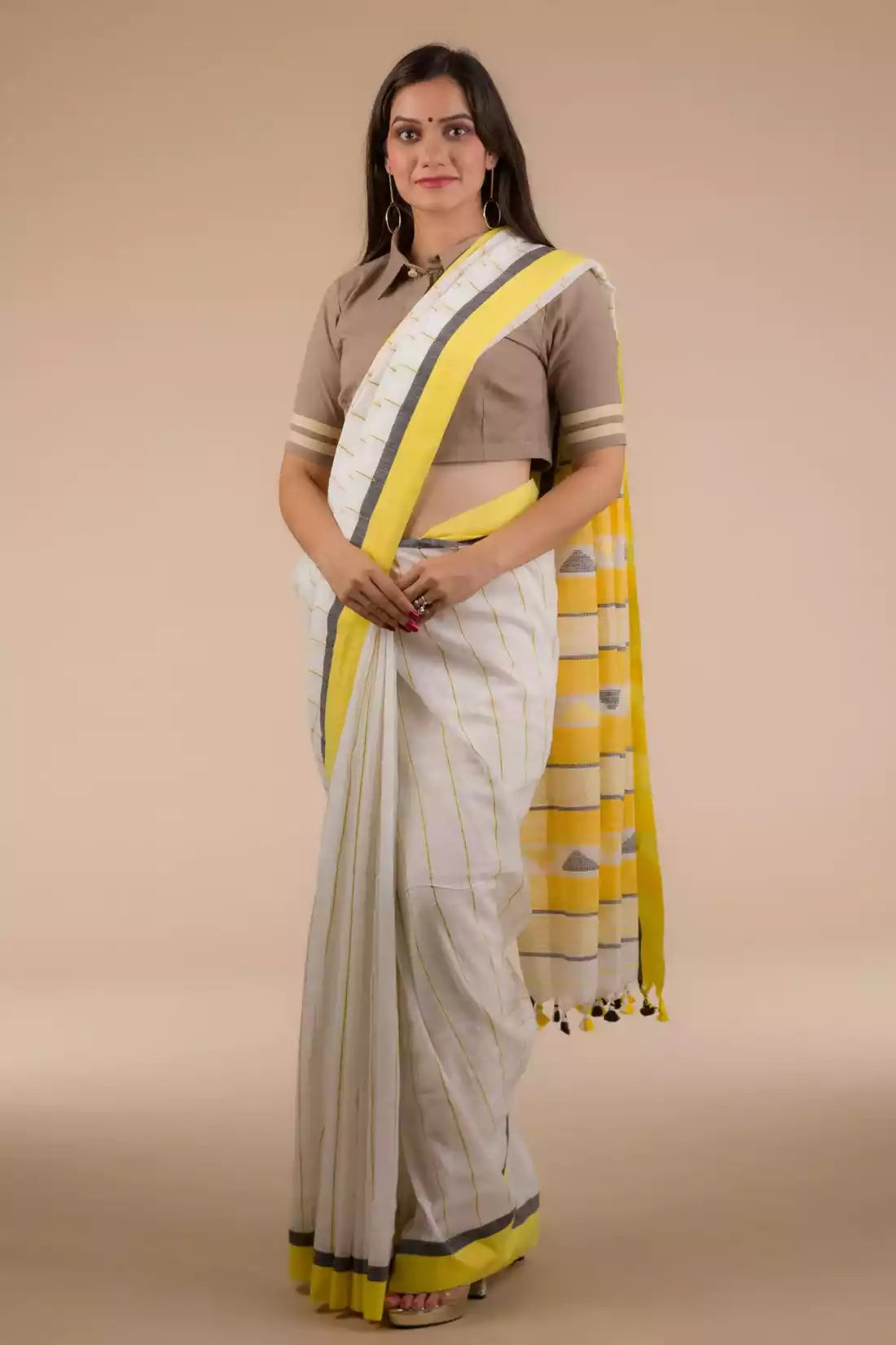 Front view of a woman in a white and yellow border saree standing in front of a beige backdrop