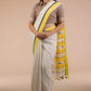 a woman in a white and yellow border saree standing in front of a beige backdrop