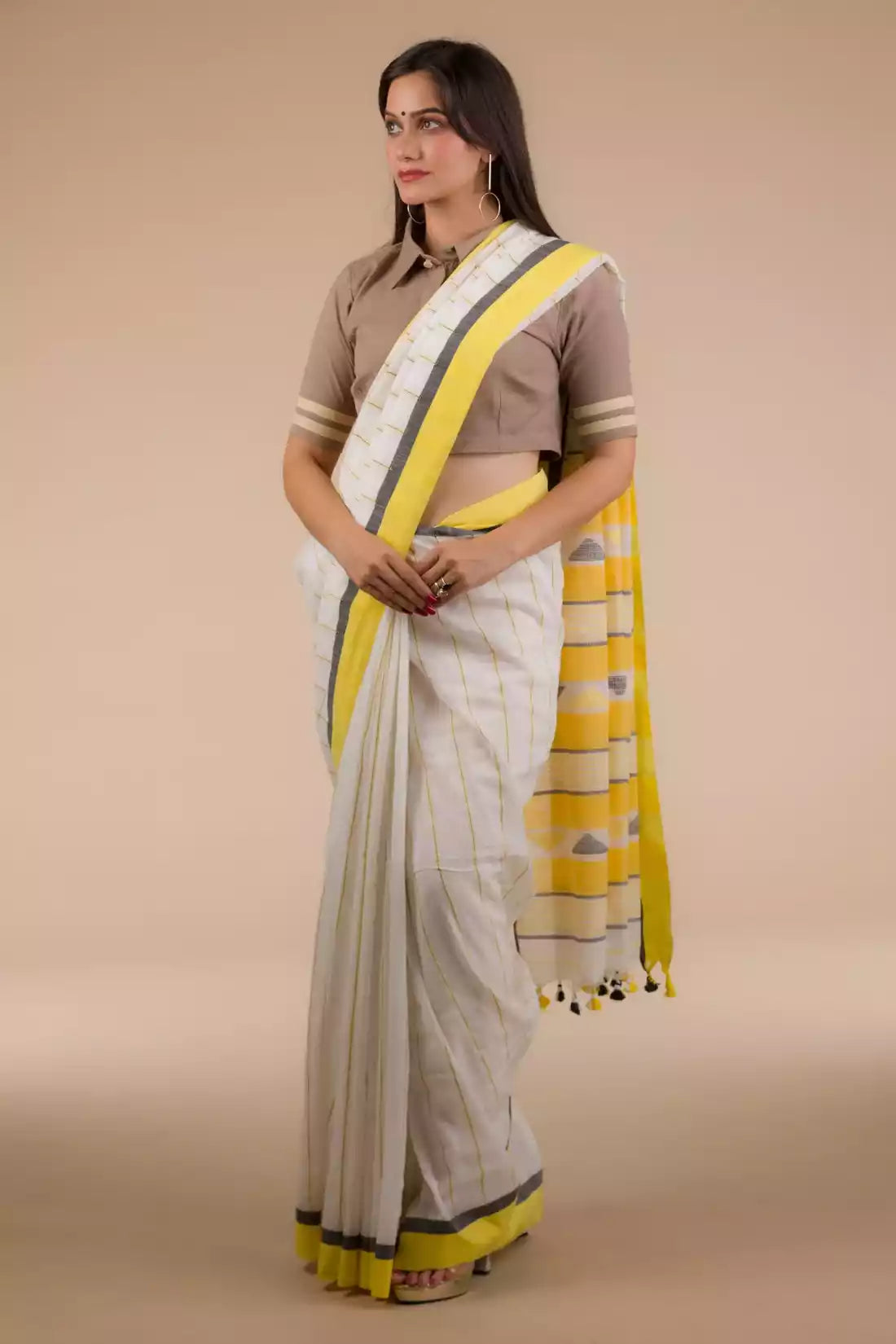a woman in a white and yellow border saree standing in front of a beige backdrop