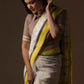 a woman in a white and yellow border saree looking down in front of a beige backdrop