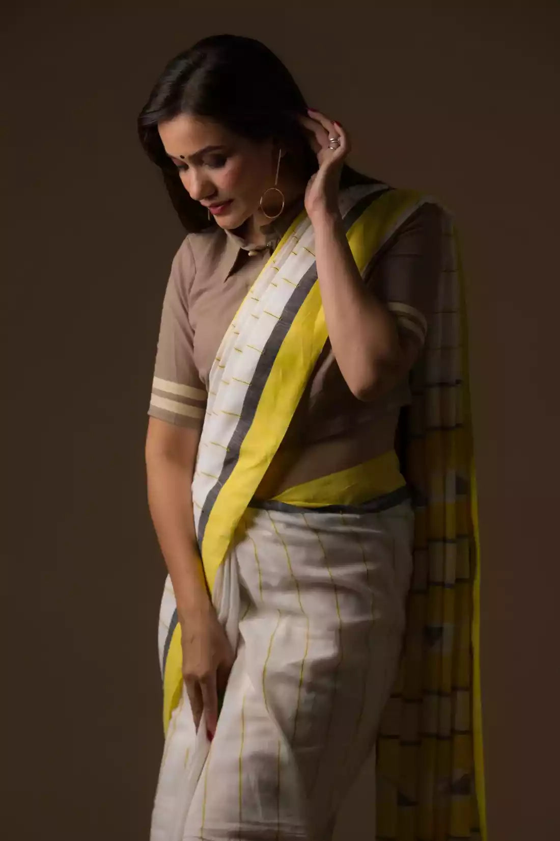 a woman in a white and yellow border saree looking down in front of a beige backdrop