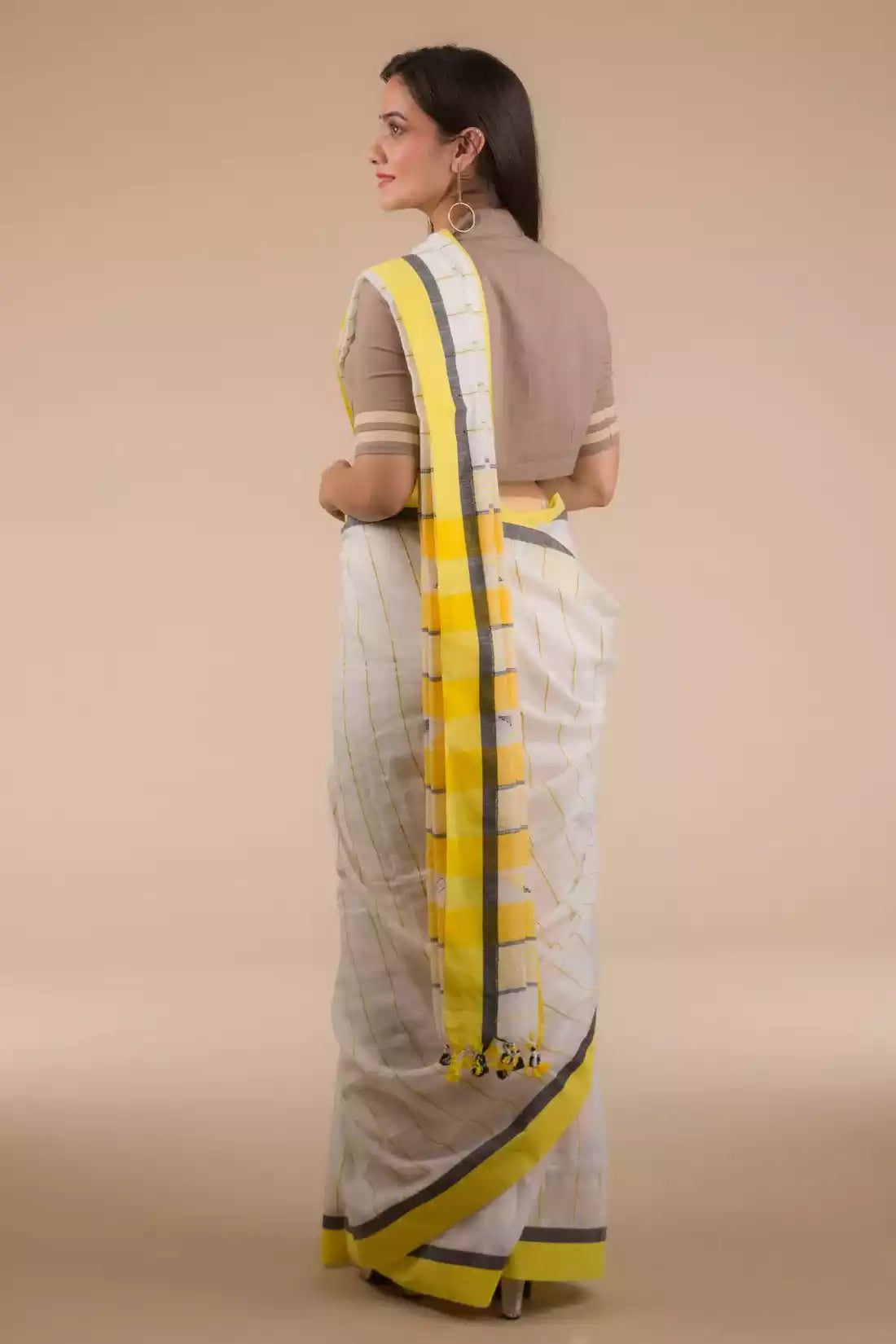 a side view of a  woman in a white and yellow border saree standing in front of a beige backdrop