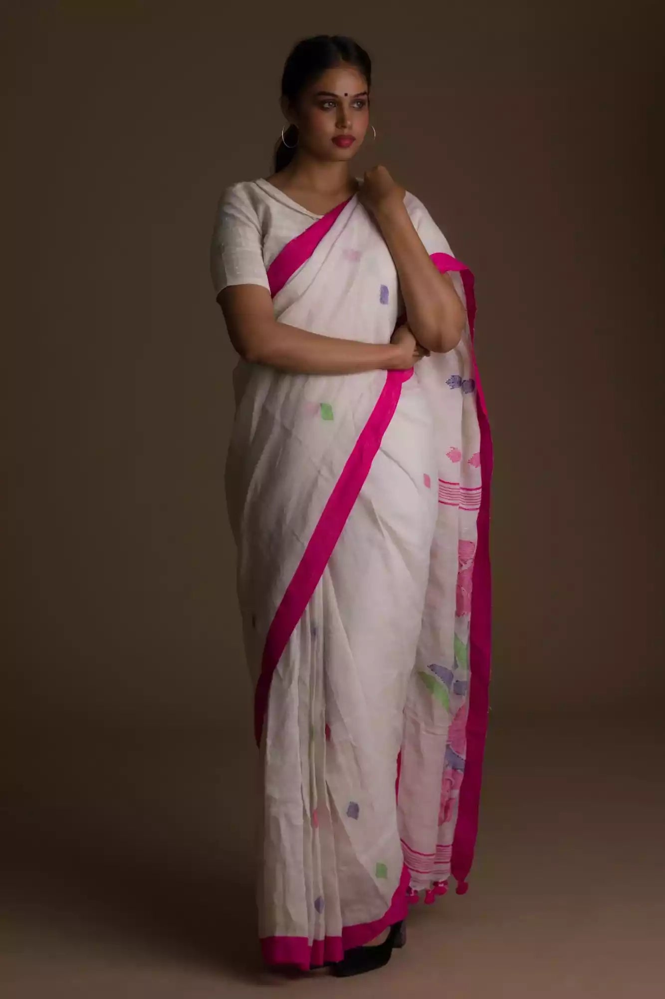 a woman standing in front of a beige backdrop wearing a white saree with pink polka dot work on it and the bottom half