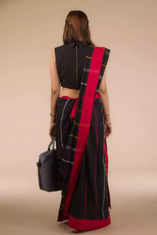 a back view of a woman standing in an ethnic black and red saree with striped fabric on the bottom and a grey bag at one side